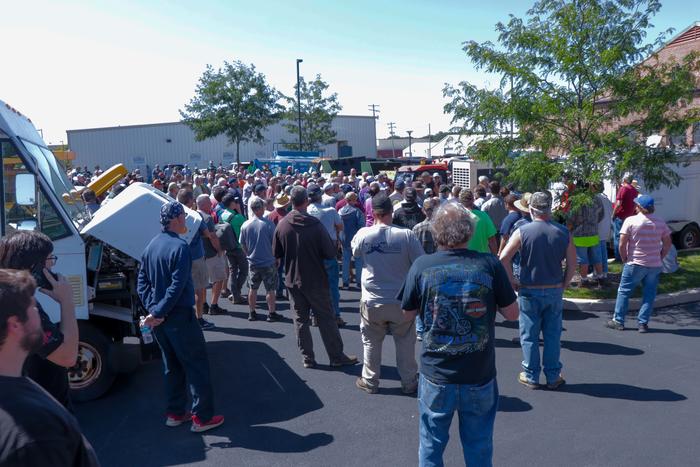 2023 Fall Auction; people standing in a crowd, staring at the auction. Truck with it's hood up to the right of the shot. Building in background.